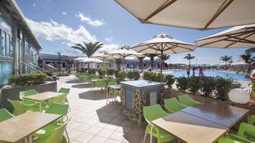 Un espace extérieur aménagé en terrasse, avec des chaises et des tables vertes, des parasols, et une piscine en arrière-plan.