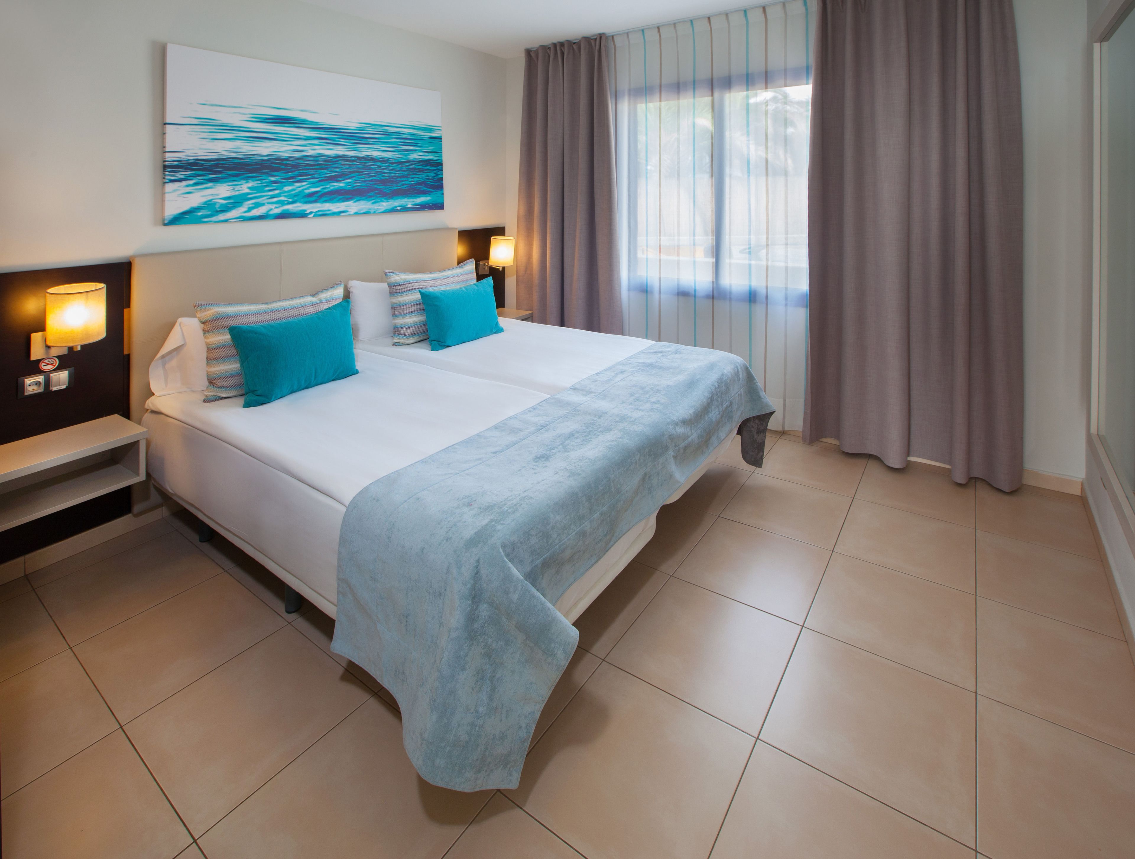 Ein Hotelzimmer mit einem großen Bett, zwei Nachttischen, einem an der Wand befestigten Kunstwerk und einem Fenster mit Vorhängen.