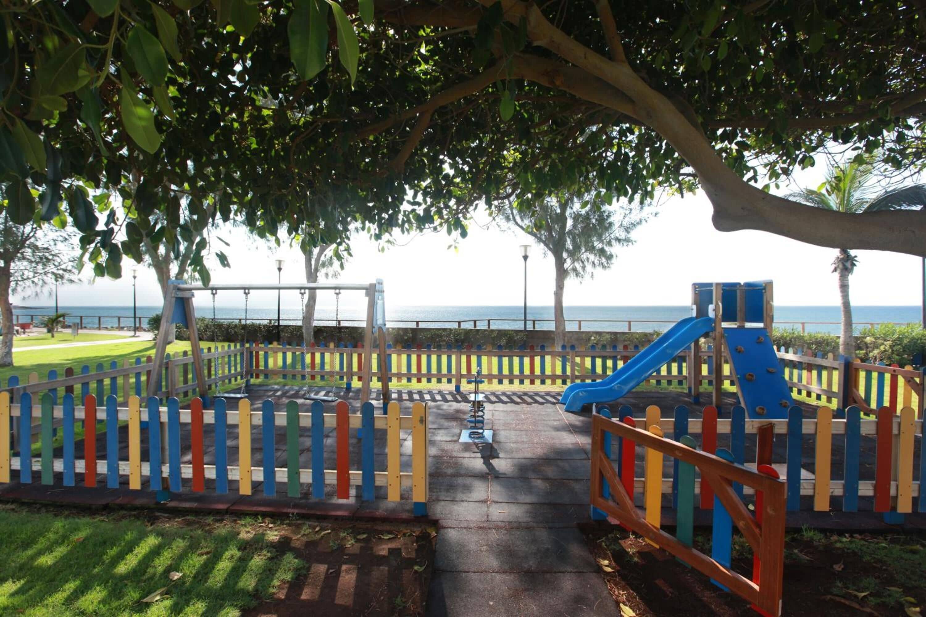 Un parque infantil con tobogán y columpios, rodeado por una valla de colores, con vista al mar al fondo.