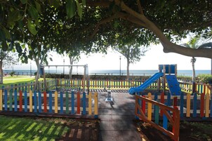 A playground with a slide and swings, surrounded by a colorful fence, with a view of the sea in the background.