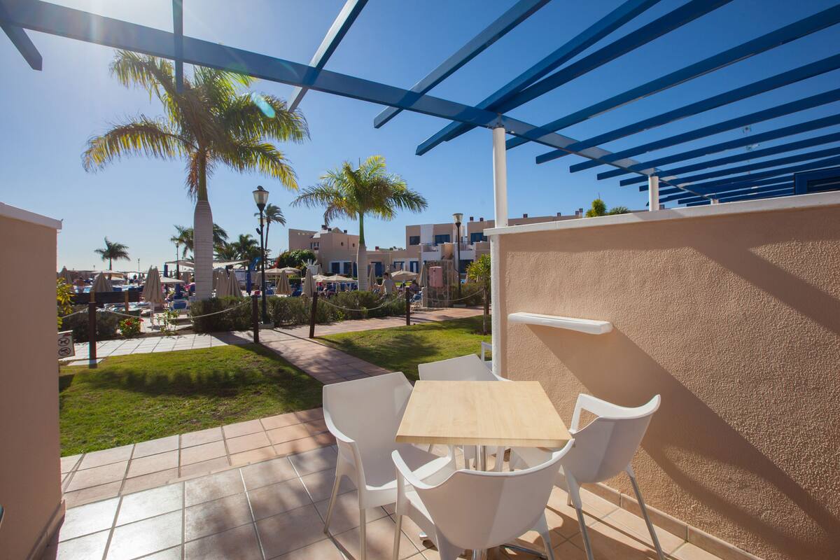 A patio with white chairs and a table, overlooking a pool and palm trees.