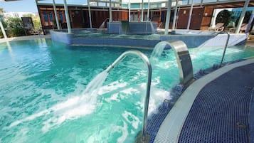 A large indoor swimming pool with clear blue water, a jacuzzi area, and a covered seating area with lounge chairs.