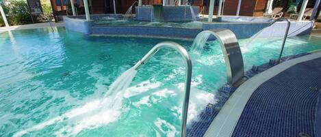 Una gran piscina interior con agua cristalina, zona de jacuzzi y área de descanso cubierta con sillones reclinables.