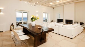 Uma sala de estar moderna com um sofá branco, uma mesa de jantar de madeira com cadeiras, uma televisão e vista para o mar através de grandes janelas.