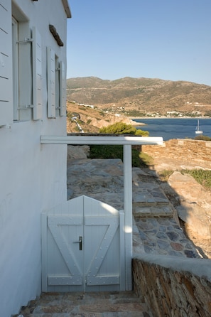 A white building with a stone staircase leading to a white door with metal studs. A stone wall and a view of the sea and hills are visible in the background.