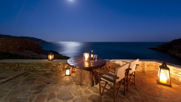 Um pátio de pedra com uma mesa redonda e cadeiras, rodeado por lanternas, com vista para o litoral à noite.