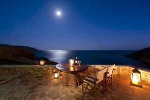 A stone patio with a round table and chairs, surrounded by lanterns, overlooking a coastal view at night. - Athina Ios Villa (Ios)