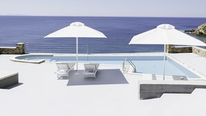 Uma área de piscina com duas guarda-sóis brancos, espreguiçadeiras e vista para o mar.