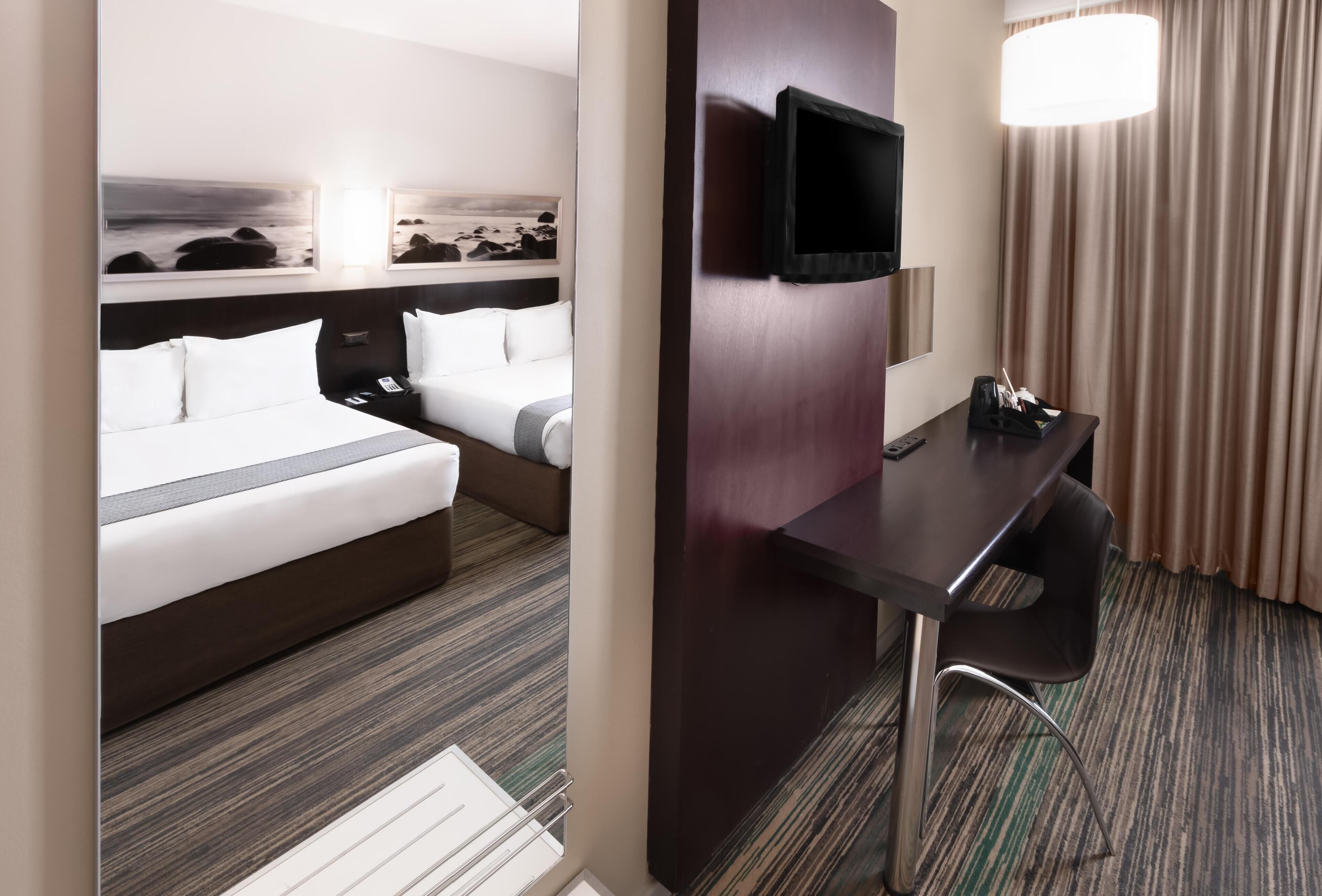 A hotel room with a bed, a desk, a chair, a television, and artwork on the wall.