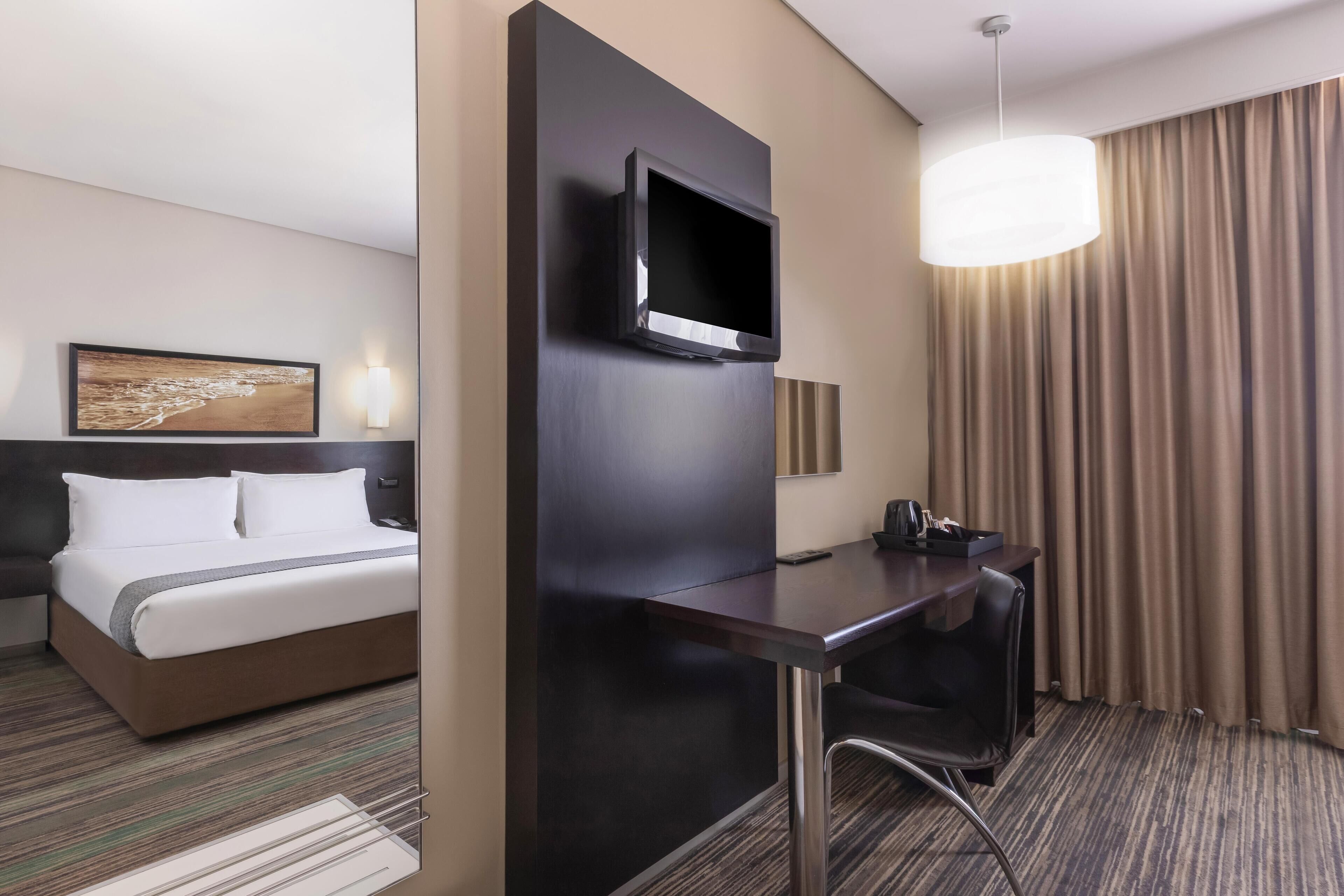 A hotel room with a bed, a desk, a TV, a lamp, and a painting on the wall.