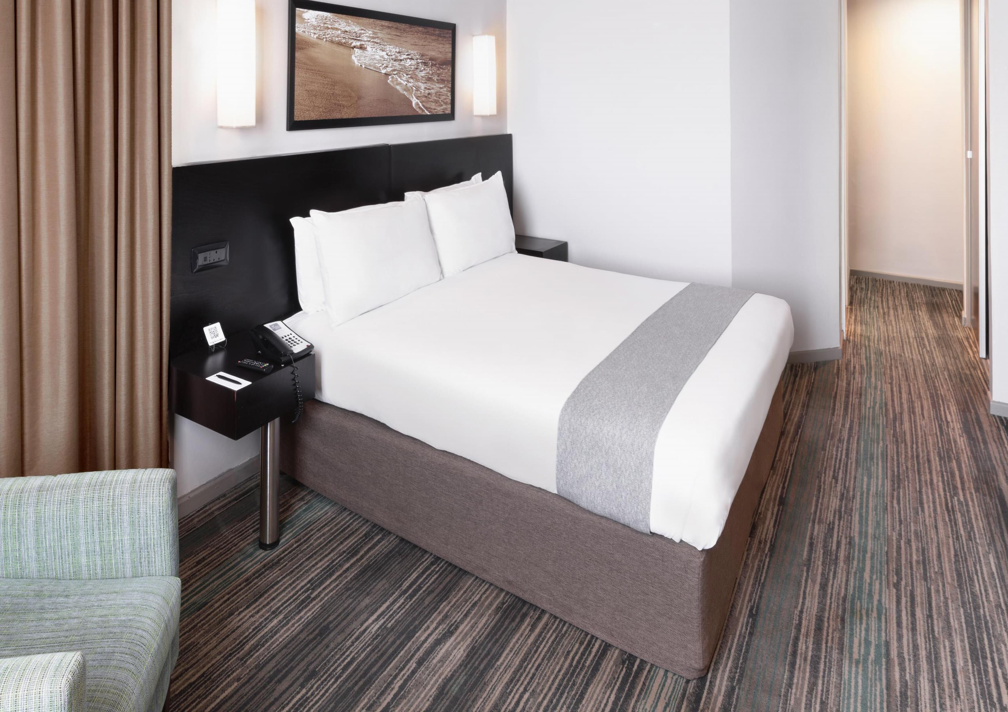 A hotel room with a large bed, a bedside table with a phone, a chair, and a picture on the wall.