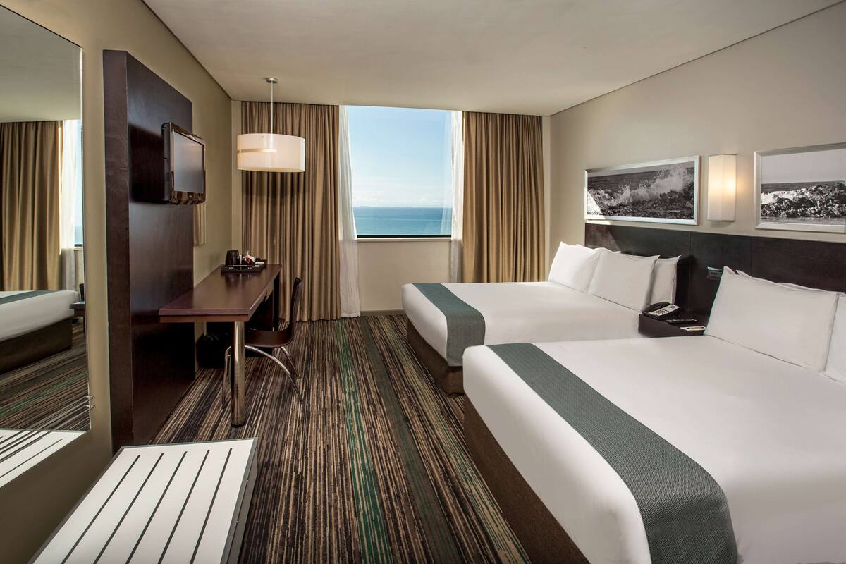 Une chambre d’hôtel avec deux lits, un bureau, une chaise, une télévision et une grande fenêtre donnant sur la mer.