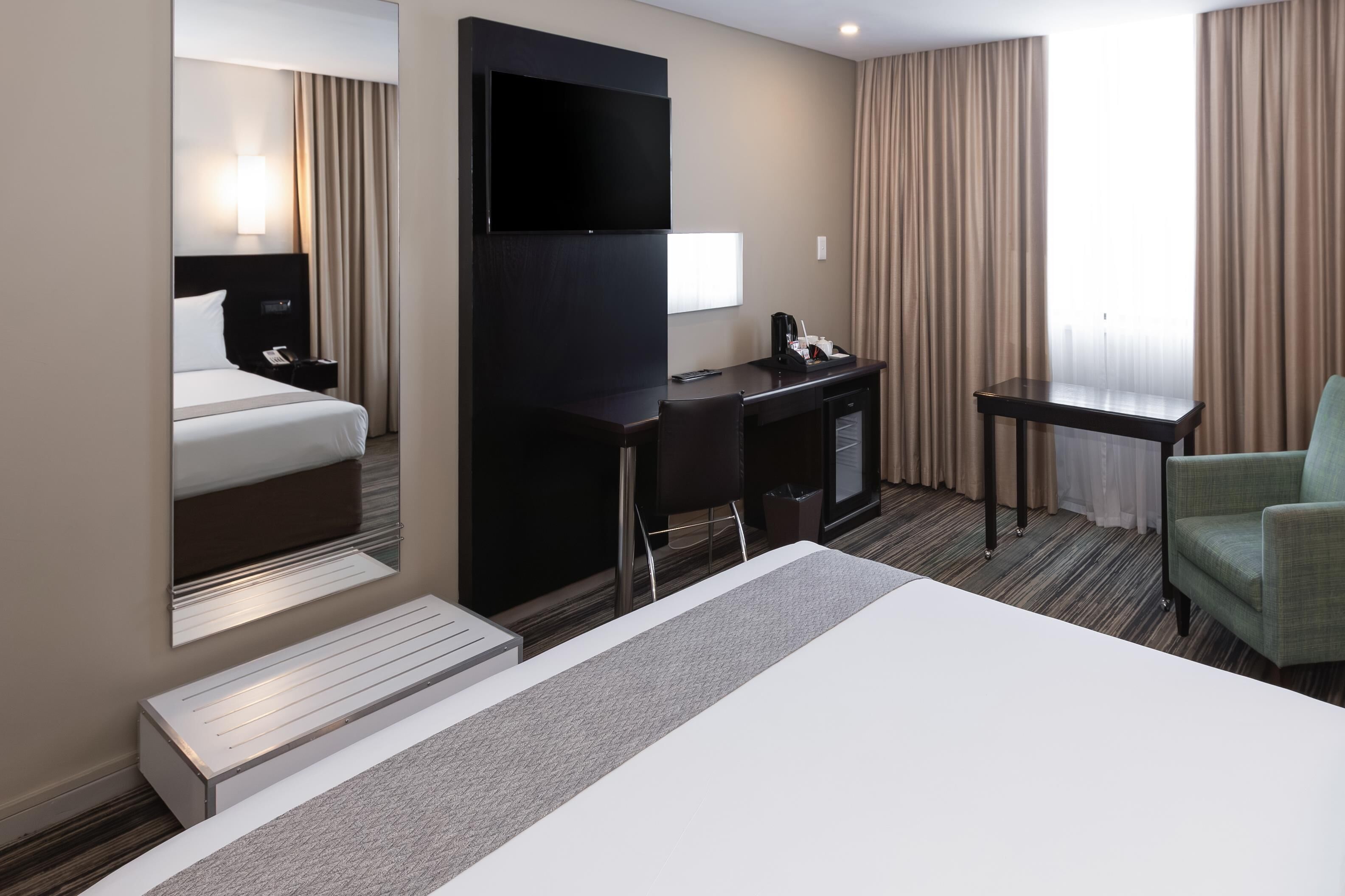 Habitación de hotel con cama, escritorio, silla, televisor y ventana con cortinas.
