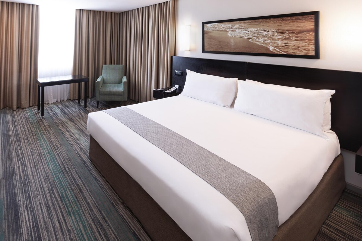 A hotel room with a large bed, a desk, a chair, and a painting on the wall.