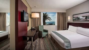 Een hotelkamer met een bed, een bureau met stoel, een televisie, een groot raam met uitzicht op de oceaan en een schilderij aan de muur.