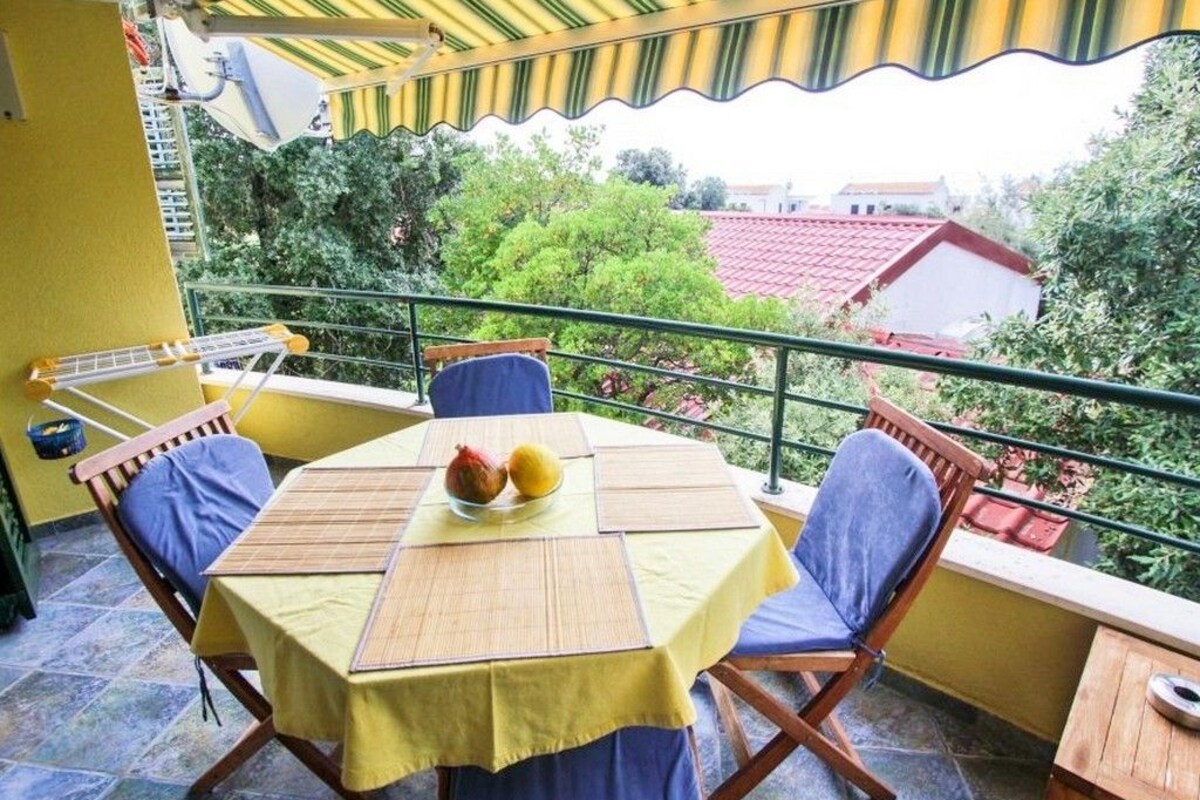 A balcony with a yellow table and blue chairs, overlooking a red-roofed building and greenery.