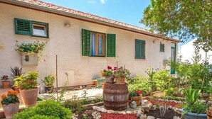 A rustic building with green shutters, a garden with various plants and a barrel, and a clear sky. - Apartments Irena (Zadar)