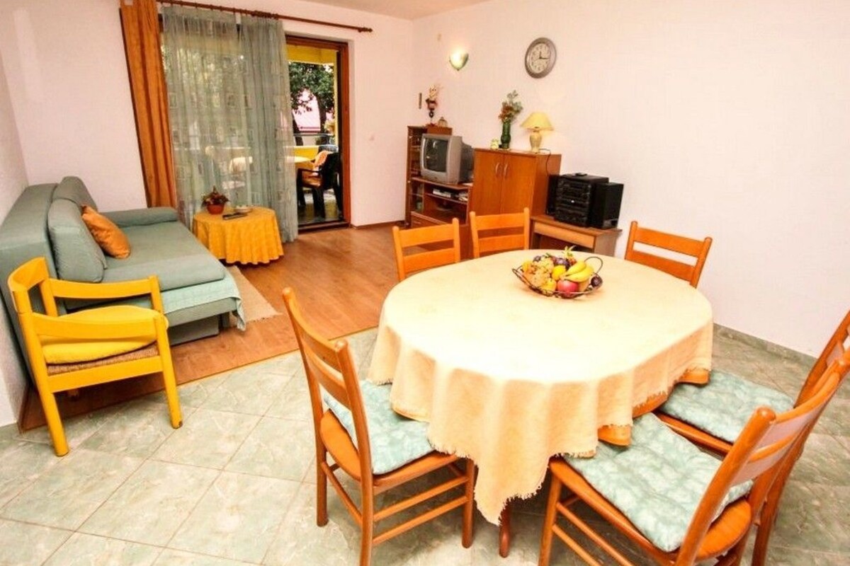 A living room with a round table, chairs, a sofa, a television, and a dining area with a fruit bowl.