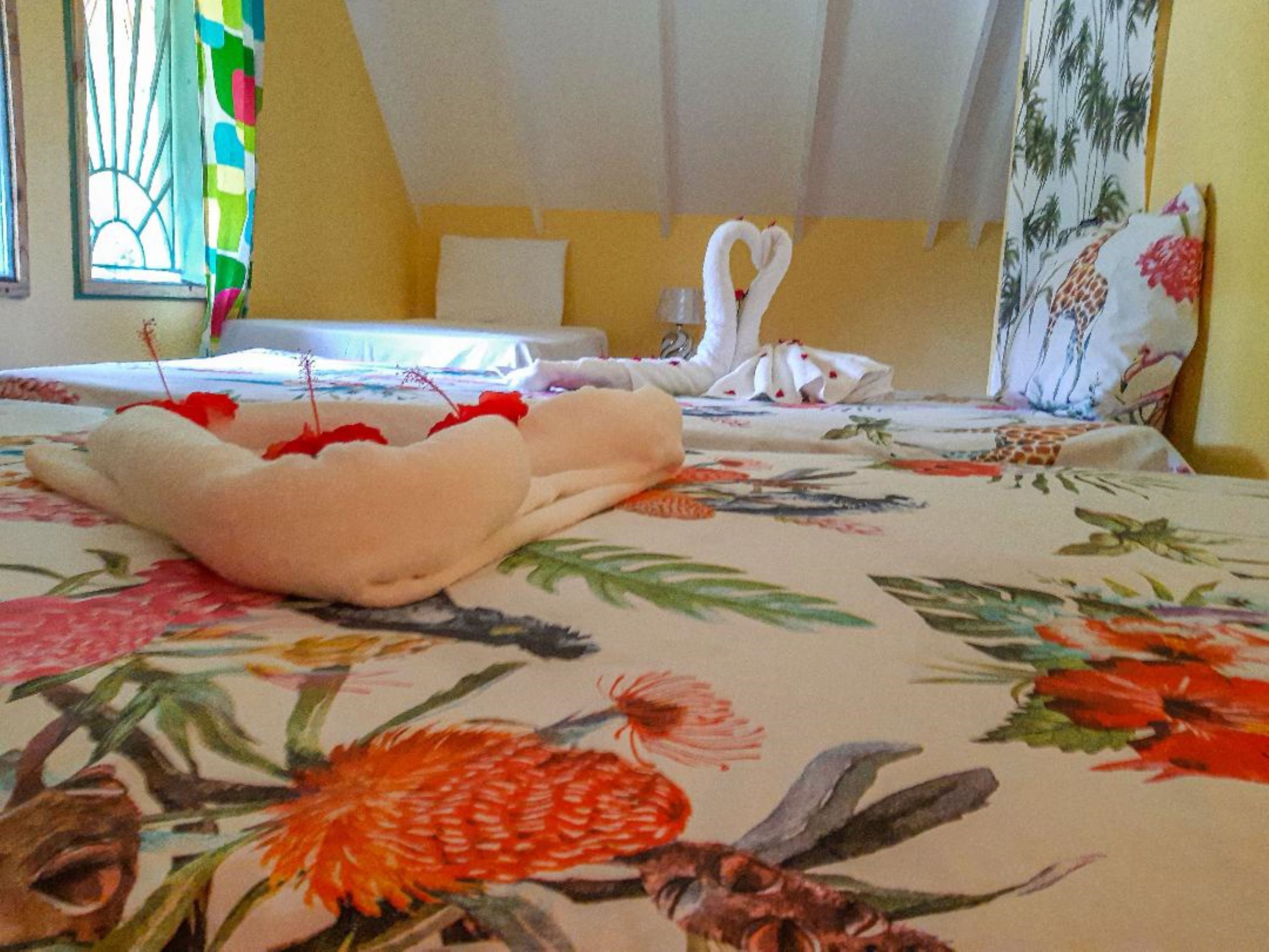 Ein Hotelzimmer mit floralem Bettüberwurf, einem Bett mit weißen Handtüchern und einer herzförmigen Dekoration, einem Fenster mit bunter Vorhang und einer Wand mit Blumenmuster.