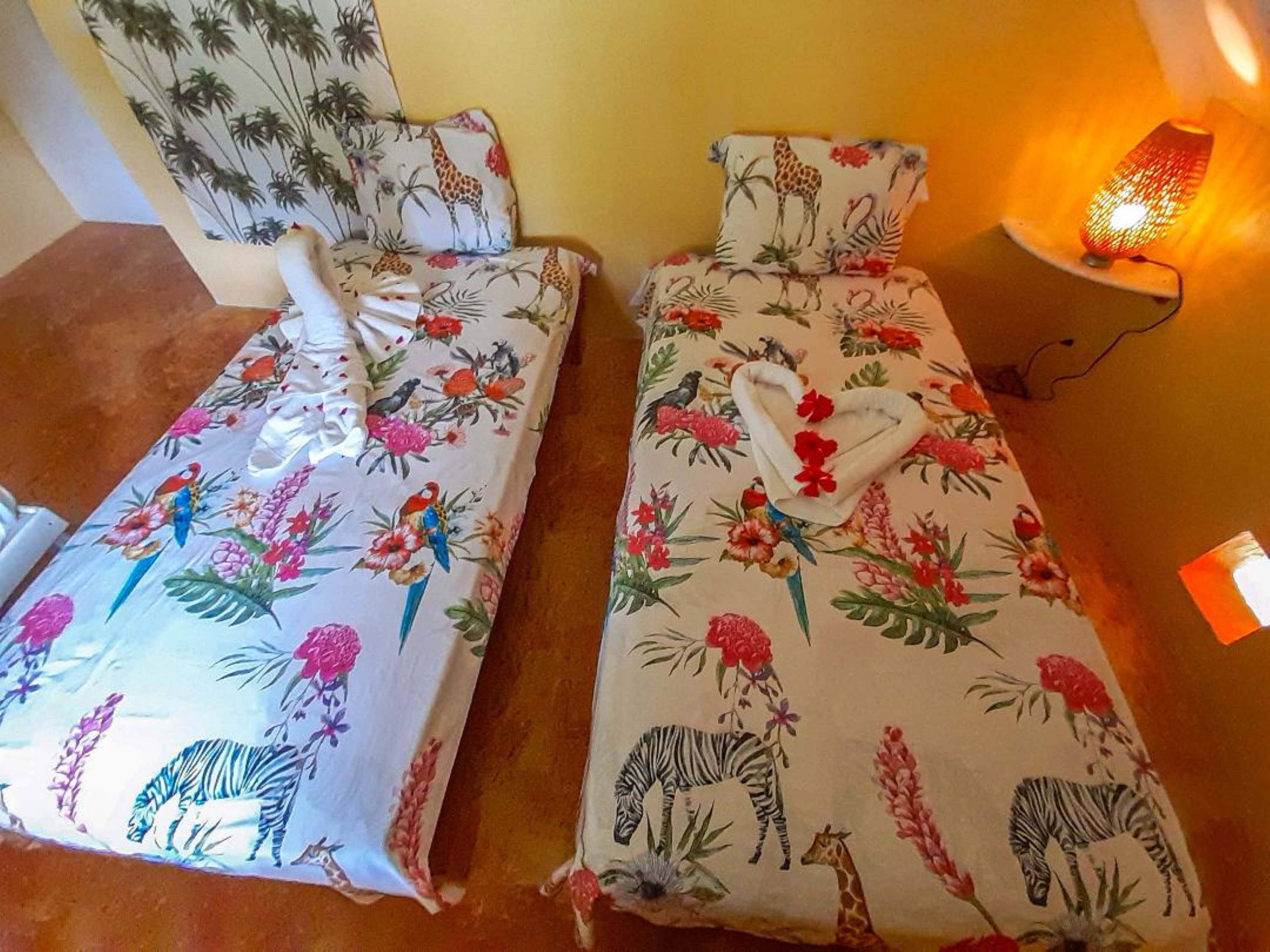 Two twin beds with floral and animal prints, a nightstand with a lamp, and a wall with a plant mural.