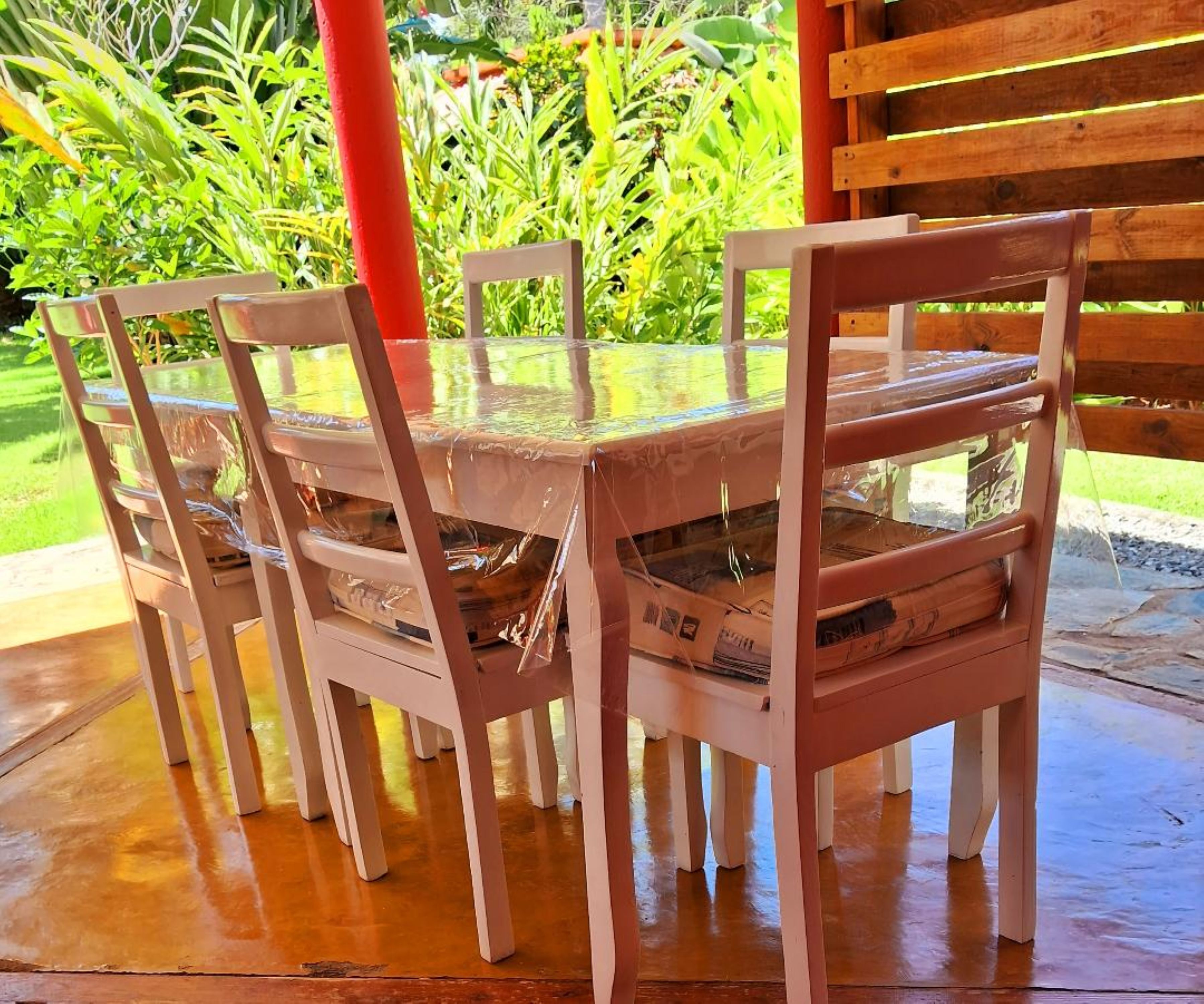 Une table ronde en verre, entourée de quatre chaises, chacune dotée d’une structure en bois et d’un siège rembourré, est placée sur une terrasse en bois, avec un jardin en arrière-plan.