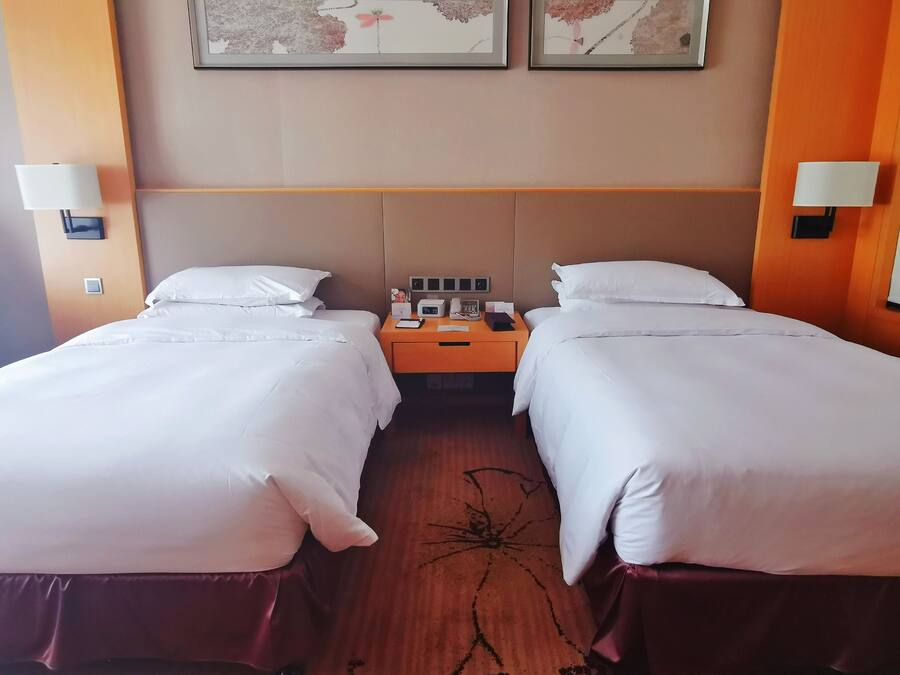 A hotel room with two beds, each with white linens and a wooden headboard. There is a nightstand between the beds with a lamp, a phone, and a clock. The walls are painted in a neutral color, and there are two framed pictures above the beds.