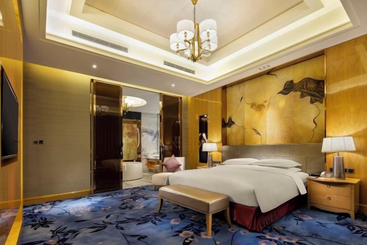 Una habitación de hotel espaciosa con una cama grande, un banco, una mesita de noche y una araña.