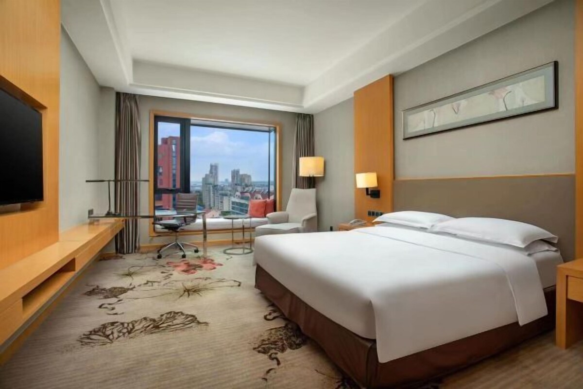 Habitación de hotel con una cama grande, un escritorio, una silla, un televisor y vistas a la ciudad.