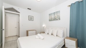 A bedroom with a bed, bedside tables, a nightstand, a wall-mounted lamp, and a door. - 335 Ocean Walk Apartments (Miami Beach)