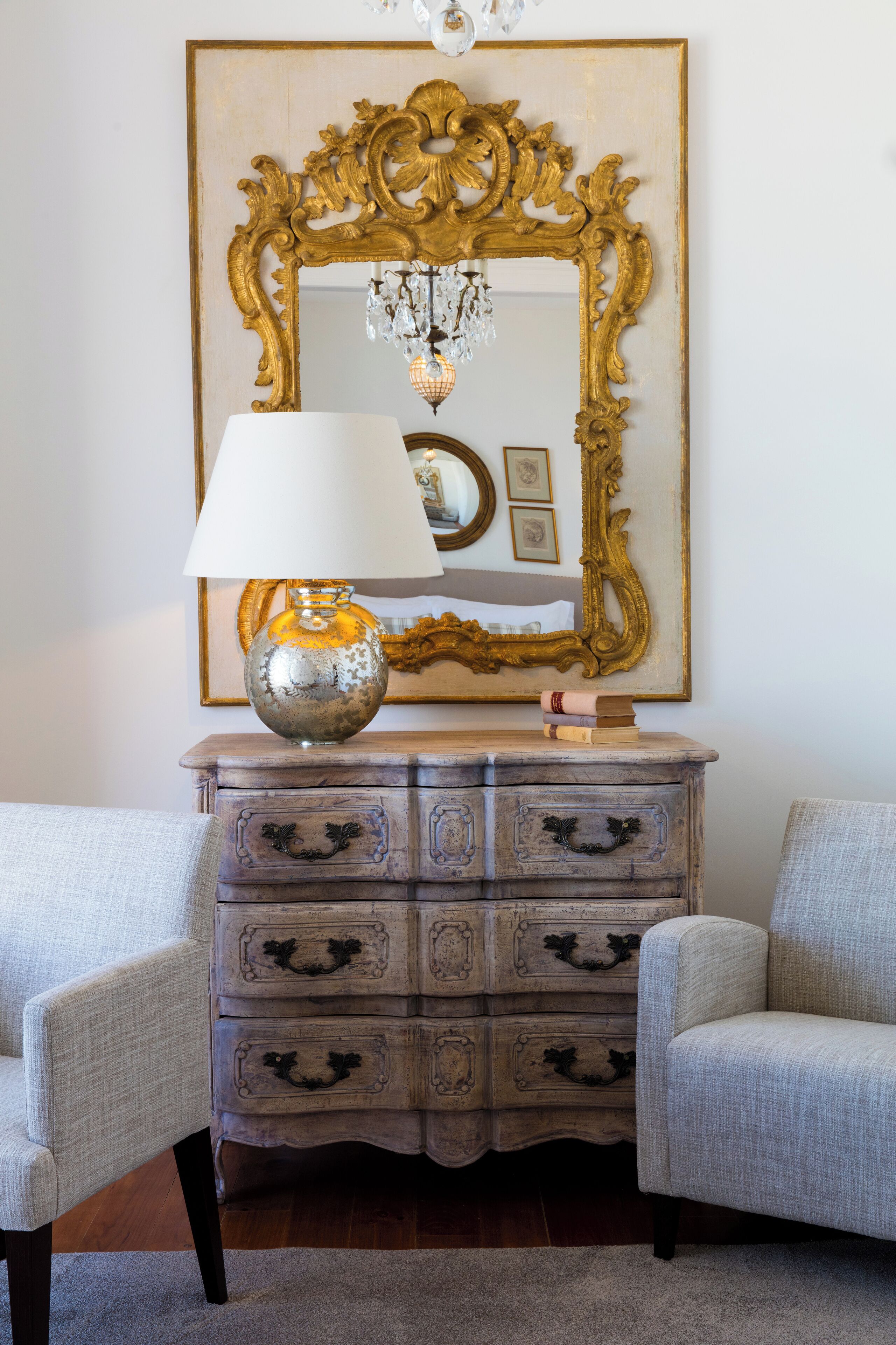 A room with a vintage dresser, a lamp, and a large ornate mirror on the wall.