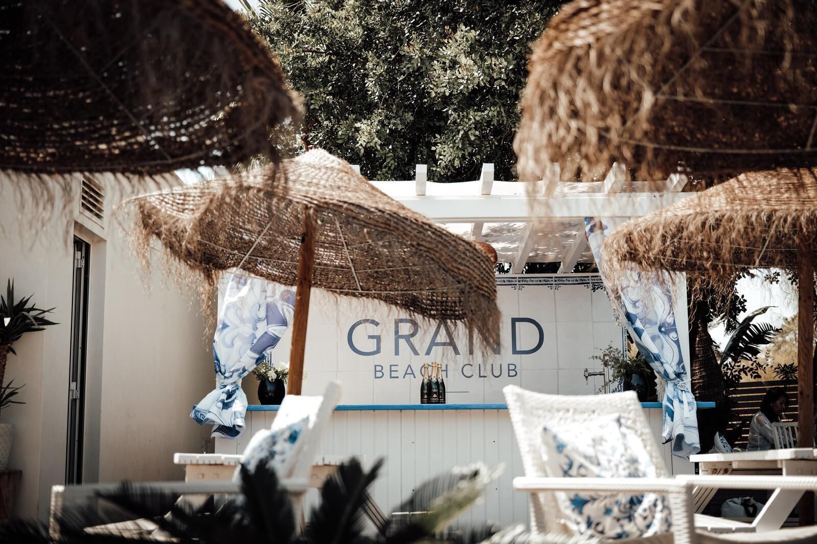 Un club de playa llamado 'GRAND' con asientos al aire libre y sombrillas de paja.
