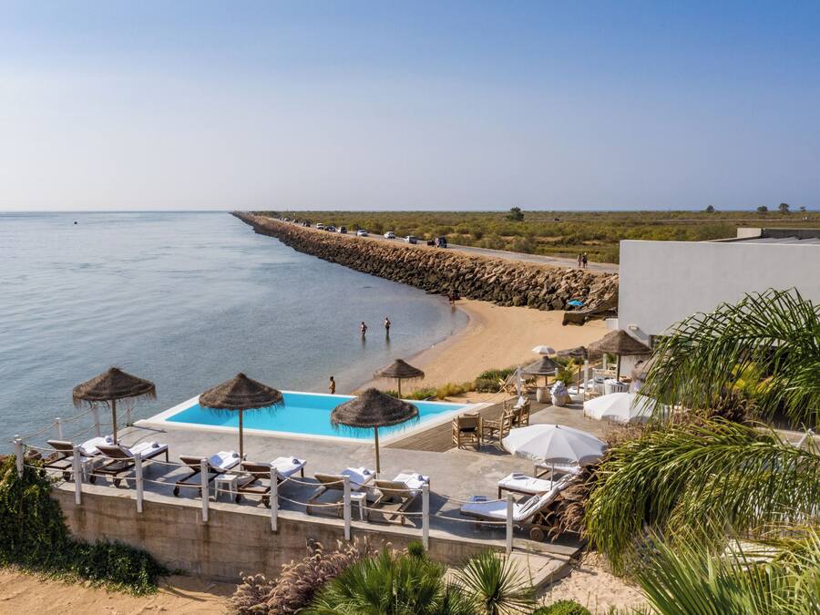 Zona de piscina con sillones de descanso y sombrillas, con vista a una playa y al mar.