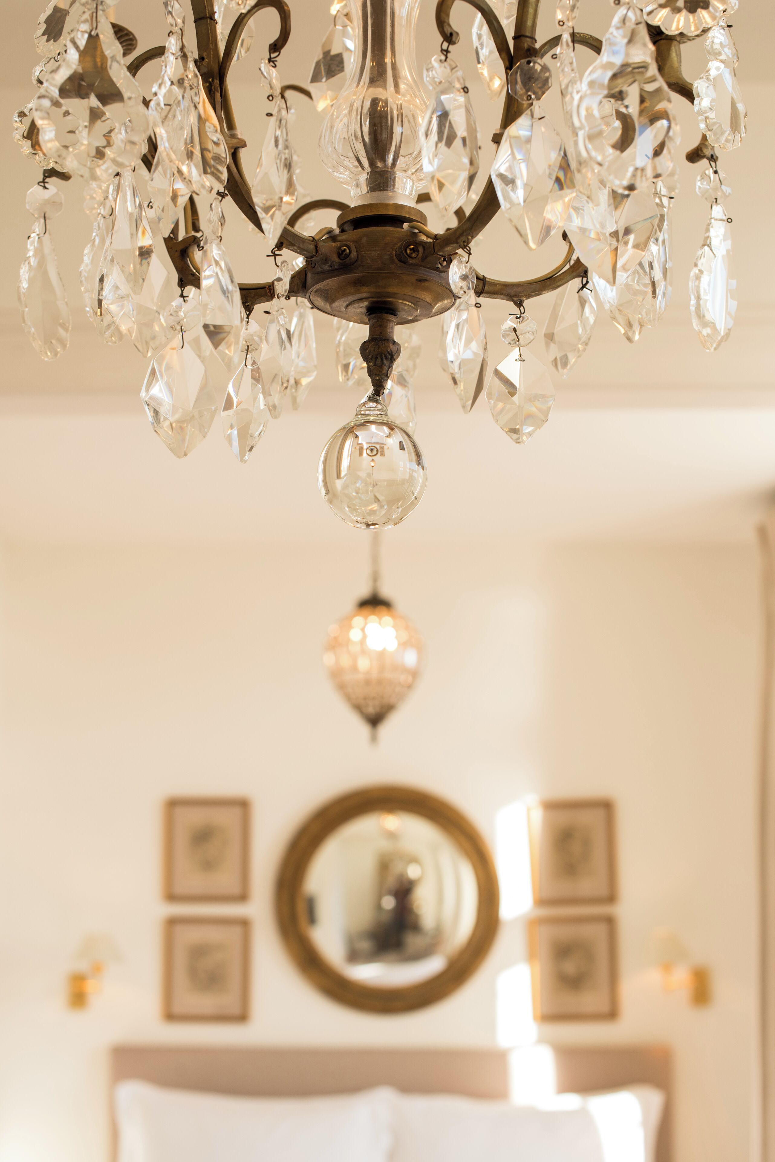 Un candelabro con múltiples elementos de cristal y una lámpara central en una habitación con cama, un espejo redondo y cuadros en la pared.