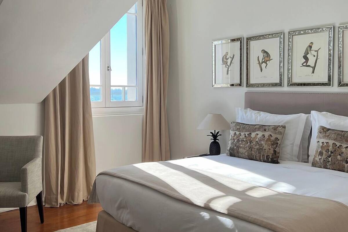 Ein Schlafzimmer mit einem großen Bett, einem Sessel und einem Fenster mit Vorhängen. An der Wand hängen drei Bilder in Rahmen.
