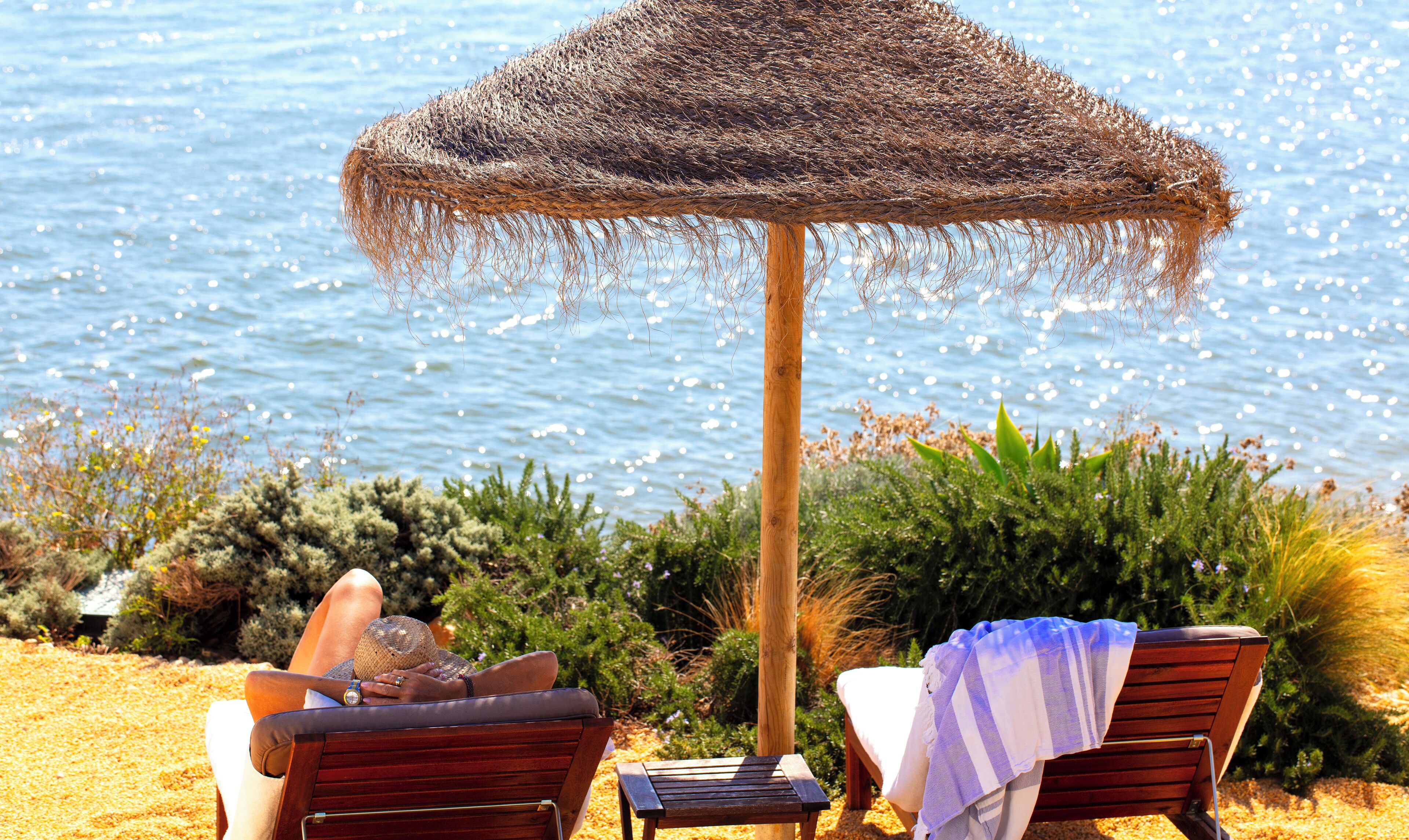 A beachside lounge area with a thatched umbrella, wooden lounge chairs, and a view of the sea.