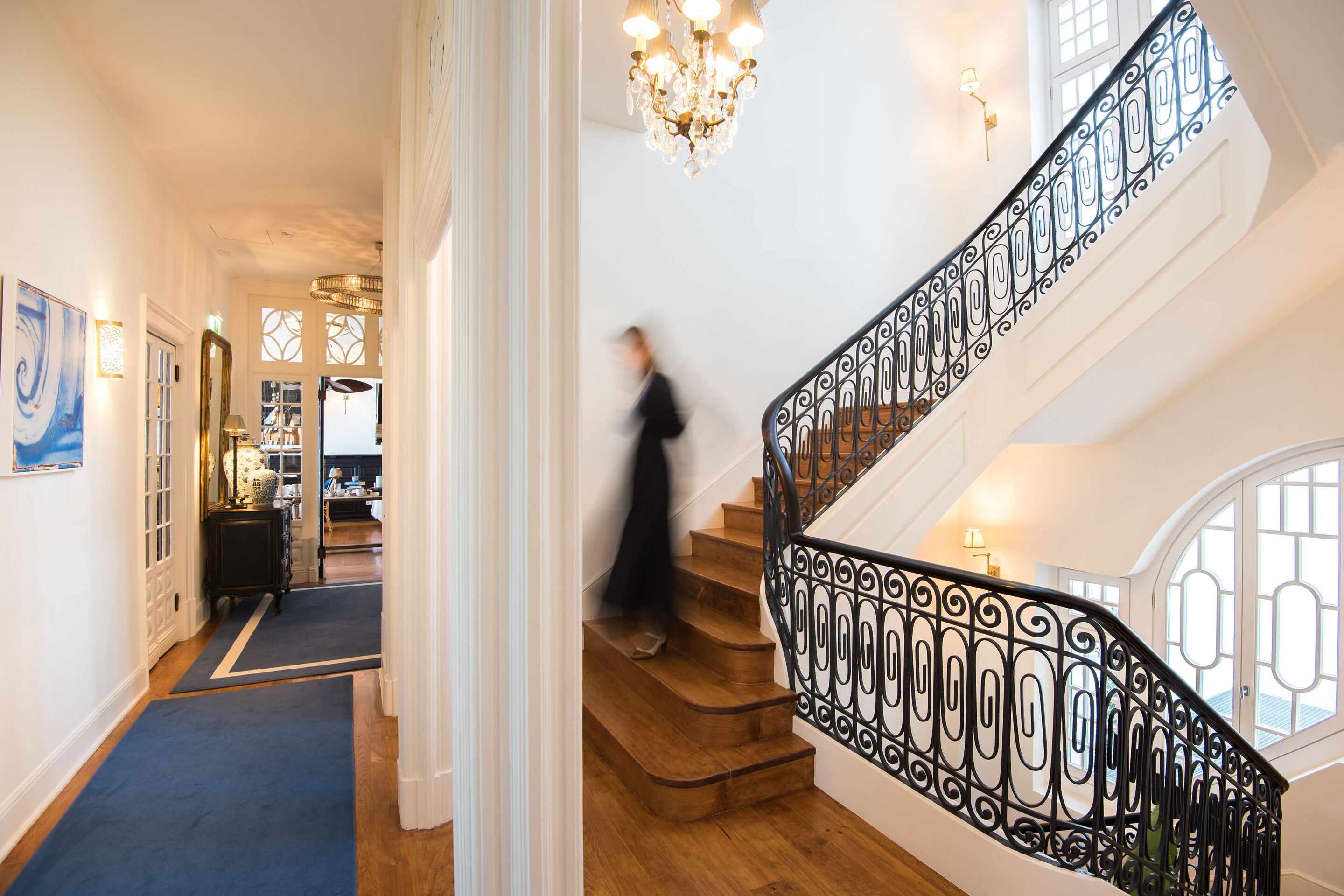 A grand staircase with ornate black railings, a chandelier, and a blue carpeted hallway leading to a room with a large window.