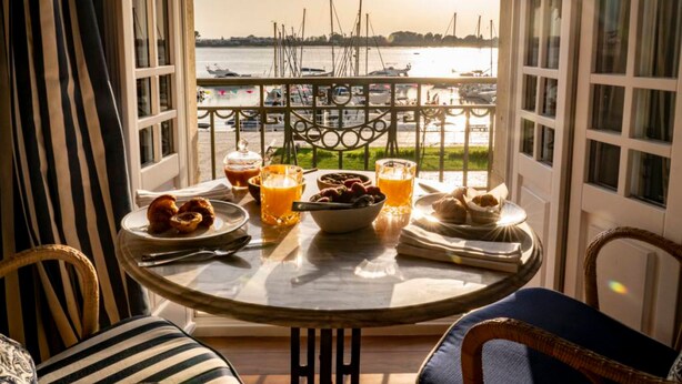 Une table à manger dressée avec des aliments et des boissons pour le petit-déjeuner, donnant sur une marina où sont amarrés des bateaux.