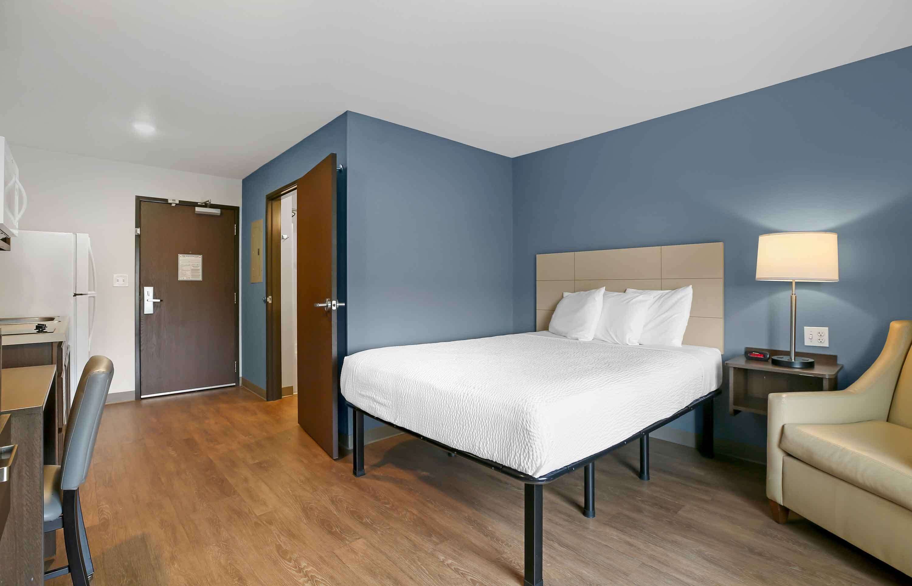 A hotel room with a bed, a desk, a chair, a lamp, and a door.