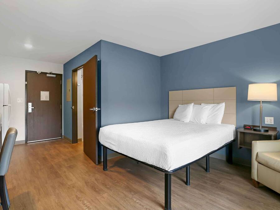 A hotel room with a bed, a desk, a chair, a lamp, and a door.