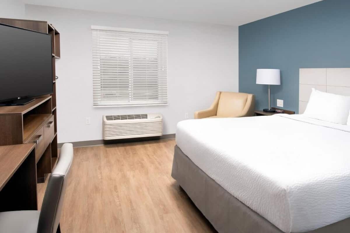 Una habitación de hotel con cama, escritorio, silla, televisión, lámpara y ventana con persianas.