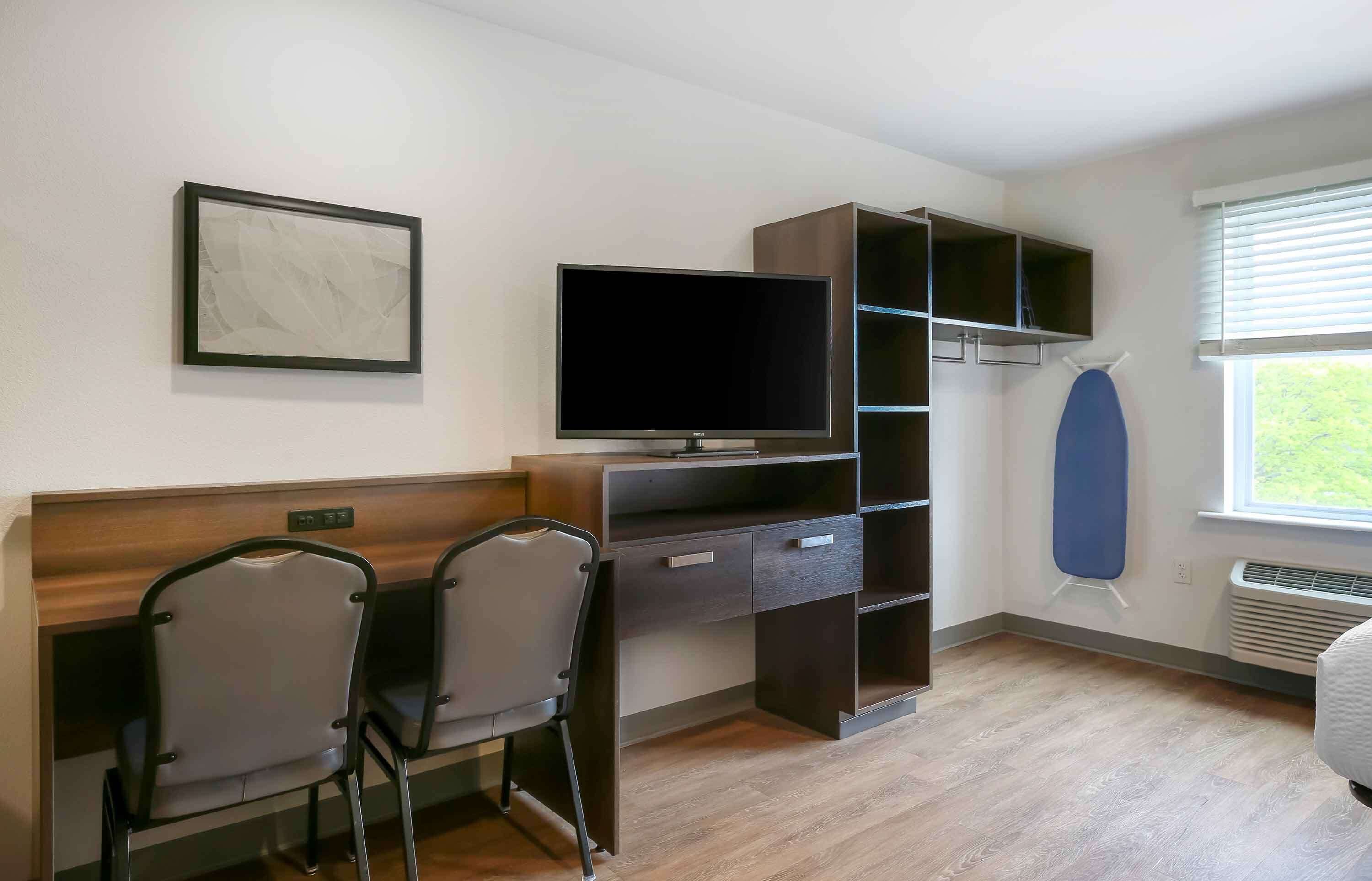 A room with a TV, two chairs, a desk, a shelf, and a window with blinds.