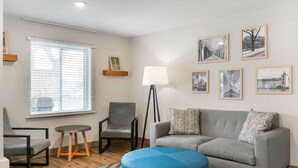 A modern living room with a grey sofa, two armchairs, a blue ottoman, a floor lamp, and a window with blinds. - Extended Stay America Suites - Providence (Providence)