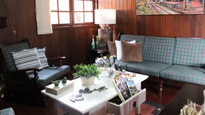 A cozy living room with wooden paneling, a plaid sofa, a matching armchair, a coffee table with magazines and a plant, and a painting of a house on the wall. - Recanto Brumas (Campos do Jordão)