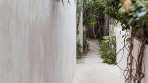 Uma trilha estreita de areia, ladeada por paredes brancas e vegetação exuberante, conduz a uma área sombreada.