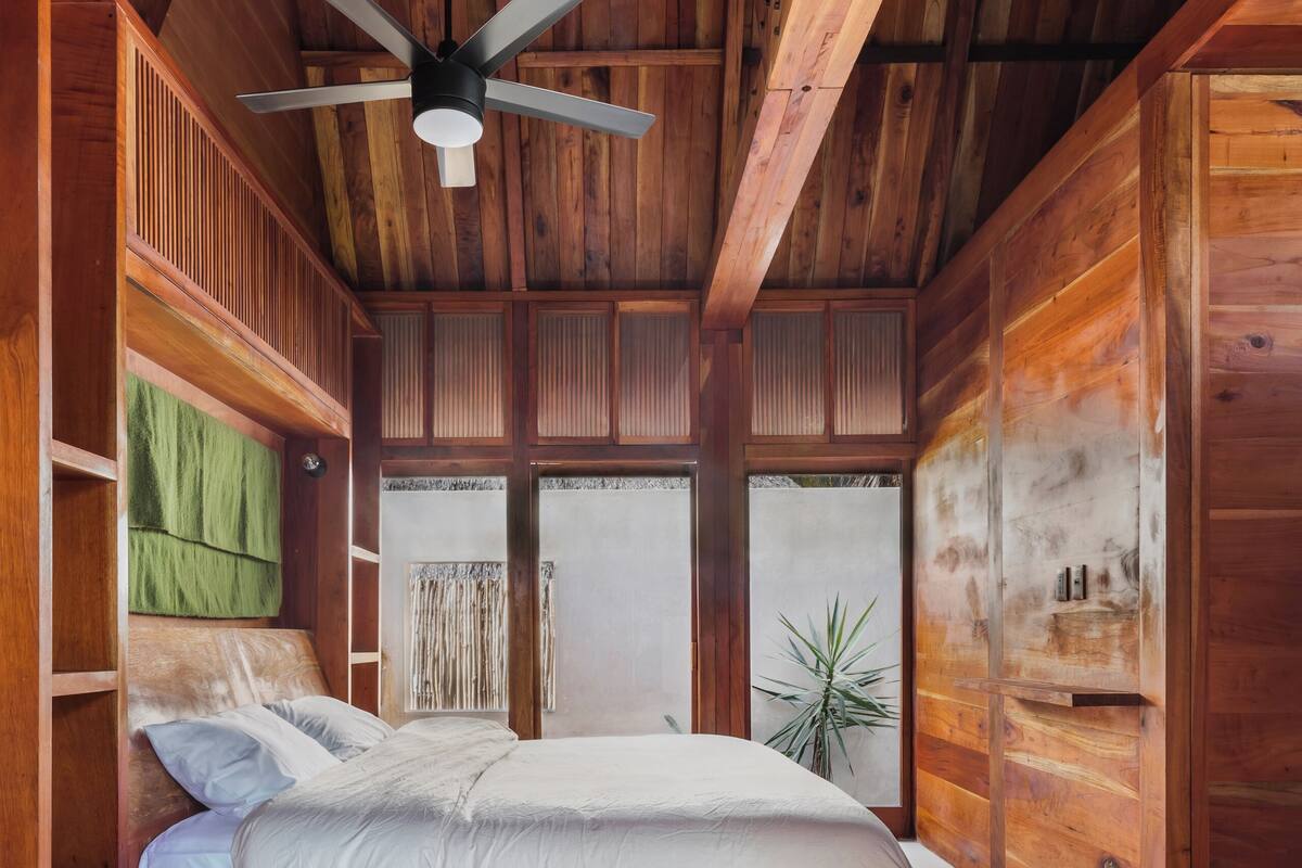 Un dormitorio con techo de madera, una cama, un ventilador de techo y una ventana con una planta.