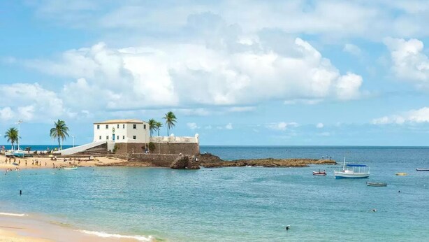 A coastal building with a staircase leading to a balcony, surrounded by palm trees and a beach. - Residencial Porto Farol (Salvador)