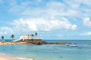 A coastal building with a staircase leading to a balcony, surrounded by palm trees and a beach.