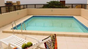 An indoor swimming pool with a tiled deck, a white lounge chair, and a few coconuts on the chair. - Residencial Porto Farol (Salvador)
