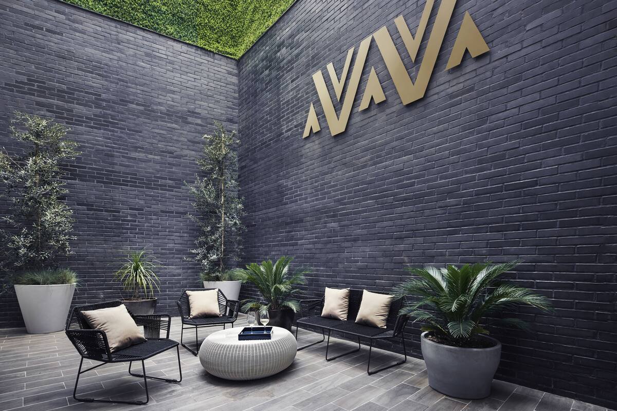 A modern outdoor seating area with black metal chairs and ottomans, surrounded by large potted plants and a dark brick wall with a large gold logo.