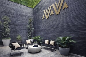 Un espace extérieur moderne, aménagé avec des chaises et des poufs en métal noir, entouré de grands pots de plantes et d’un mur de briques sombres orné d’un grand logo doré.